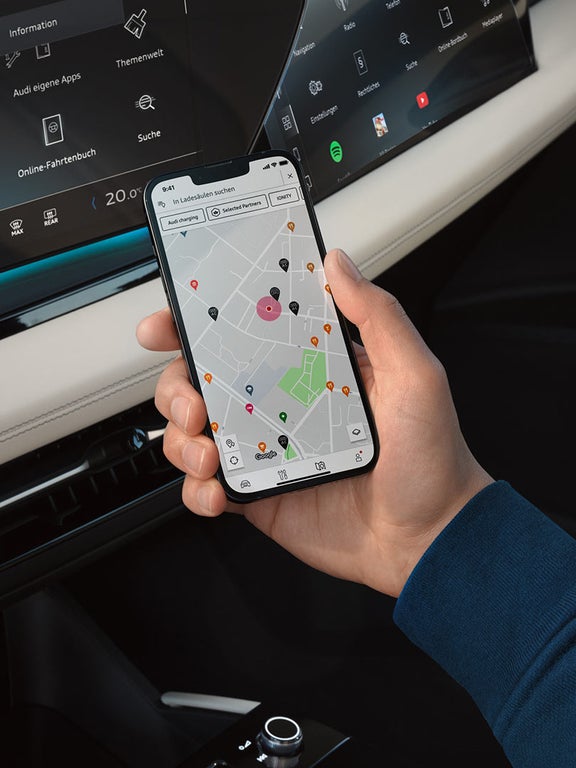 Hand hält Smartphone mit Ladesäulenkarte und Standorten, im Hintergrund Display der Audi Fahrzeugkonsole.
