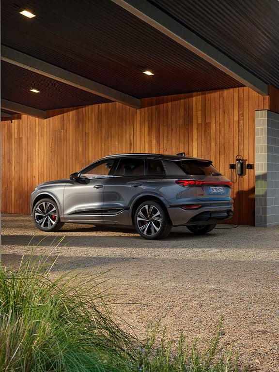 Grauer Audi SQ6 SUV e-tron lädt an Wandladestation vor Holzfassade, Fahrzeugansicht von hinten rechts.
