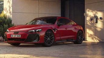Ein roter Audi e-tron GT quattro steht im Licht eines Sonnenuntergangs auf der gepflasterten Auffahrt vor einem Haus mit moderner Architektur und wird an einer Wallbox geladen. Ein Mann nähert sich dem Fahrzeug.