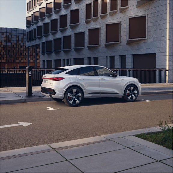 A white SUV parked at an electric charging station near modern architecture, with a sunlit urban backdrop.