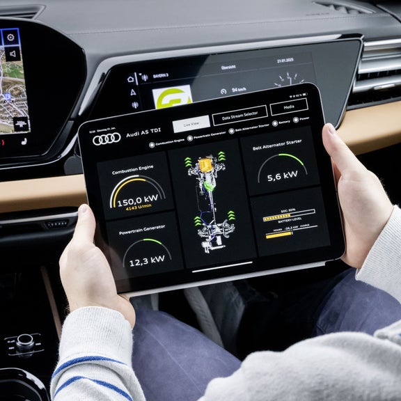 Ein Fahrer hält ein Tablet in einem Audi A5 TDI. Auf dem Bildschirm wird eine Echtzeit-Diagnose des Fahrzeugs angezeigt, inklusive Daten zu Verbrennungsmotor, Generator und Batterie. Im Hintergrund ist das Armaturenbrett mit einem digitalen Display und Navigationssystem zu sehen.