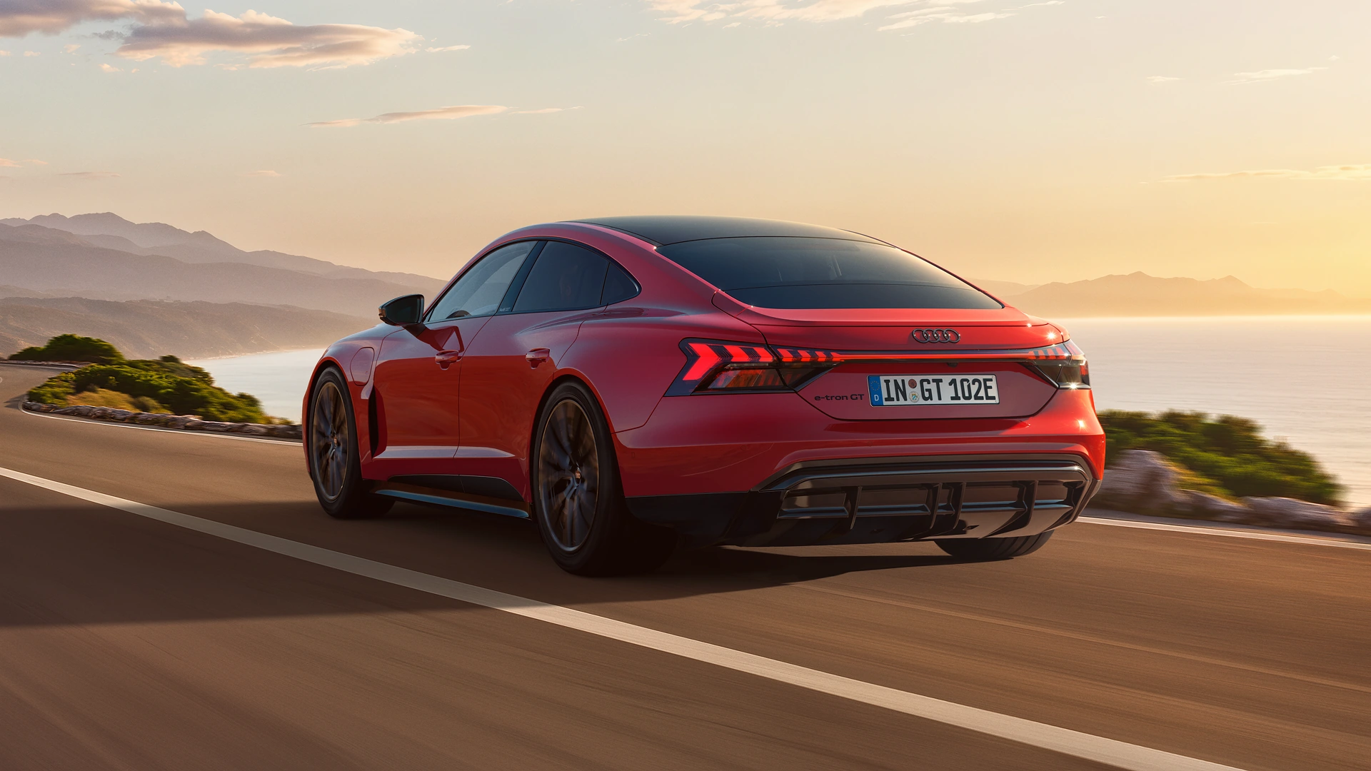 Rear three-quarter view of a red Audi e-tron GT driving on a coastal road with mountains and the sea in the background.