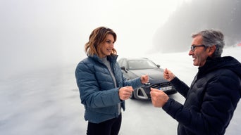 Zwei Personen (eine jüngere Frau und ein älterer Mann mit Brille) in Winterkleidung stehen auf einer Eisfläche und simulieren Fahrbewegungen, während im Hintergrund ein Audi zu sehen ist.