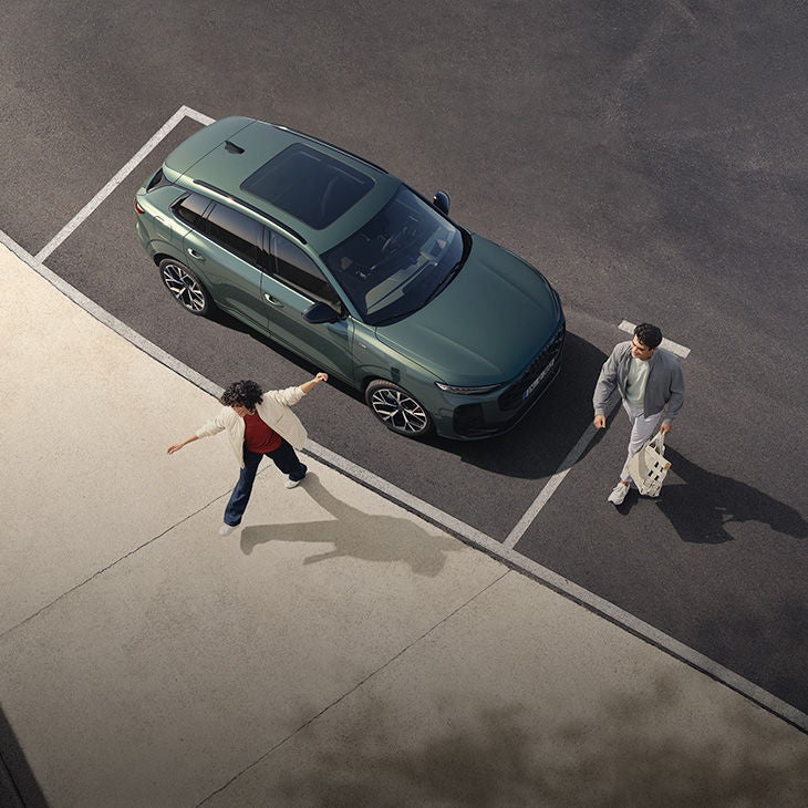 Von oben gesehen, stehen zwei Personen auf einem Parkplatz. Eine Frau, die Arme ausgestreckt, wendet sich einem Mann zu, der mit einer Tasche in der Hand geht. Ein dunkelgrüner Audi Q3 ist neben ihnen geparkt.