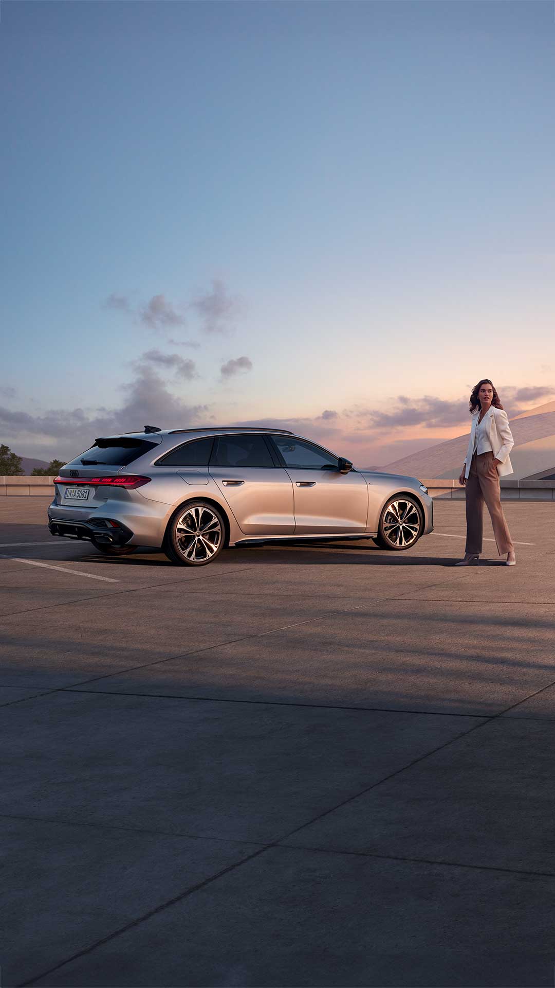 Eine Frau steht neben einem silbernen Audi Modell bei Sonnenuntergang und guckt in die Ferne
