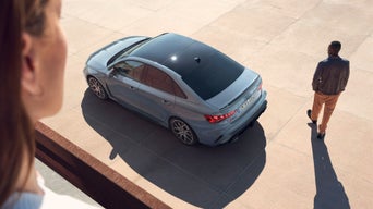 Frau im sommerlichen Outfit schiebt ihr Fahrrad an einem geparkten grauen Audi vor einem Straßencafé in sonniger Innenstadt entlang.