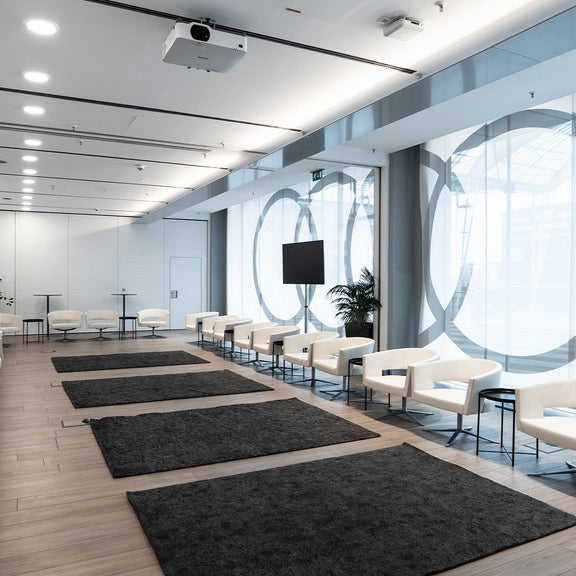 Heller Veranstaltungsraum mit Reihen weißer Sessel, Teppichen und großer Glasfront.