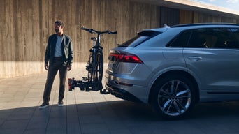 Un homme se tient derrière une Audi sur l'attelage de laquelle est installé un porte-vélos, et il porte un vélo.