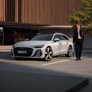 Vit Audi A6 e-hybrid parkerad mot en träbyggnad, bakom går en person.