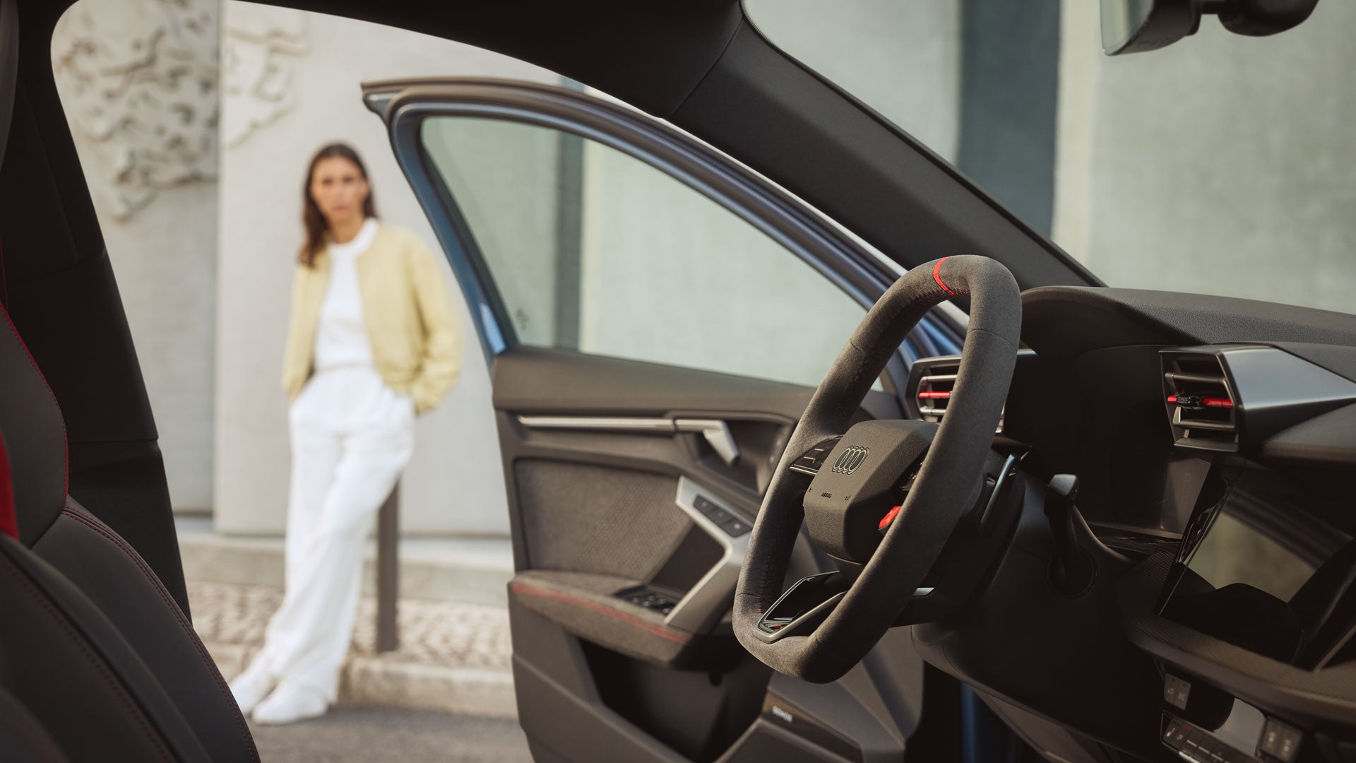Blick auf das Lenkrad und Interieur eines Audi durch die geöffnete Fahrertür, im Hintergrund steht eine Frau an einer Hauswand.