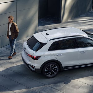 Ein Mann geht neben einem weißen Audi A3 auf einer gepflasterten Auffahrt mit moderner Gebäudefassade entlang.