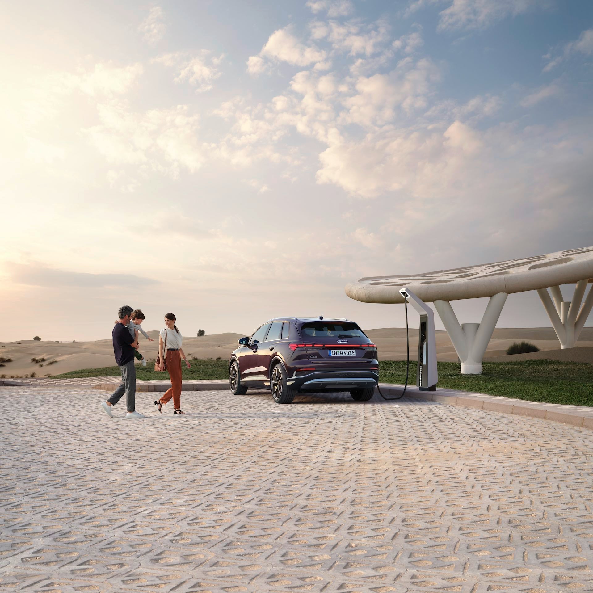 Drei Personen gehen an einem ladenden Audi Q4 e-tron vorbei, im Hintergrund Wüste, modernes Dach und Himmel mit leichter Bewölkung.