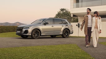 Das Bild zeigt einen silbernen Audi Q7 SUV TFIS e, der an eine Ladestation angeschlossen ist, die an der Wand eines modernen Hauses angebracht ist. Ein Mann und eine Frau gehen vom Auto weg und suggerieren damit die einfache Nutzung und Integration von Elektrofahrzeugen in das tägliche Leben.