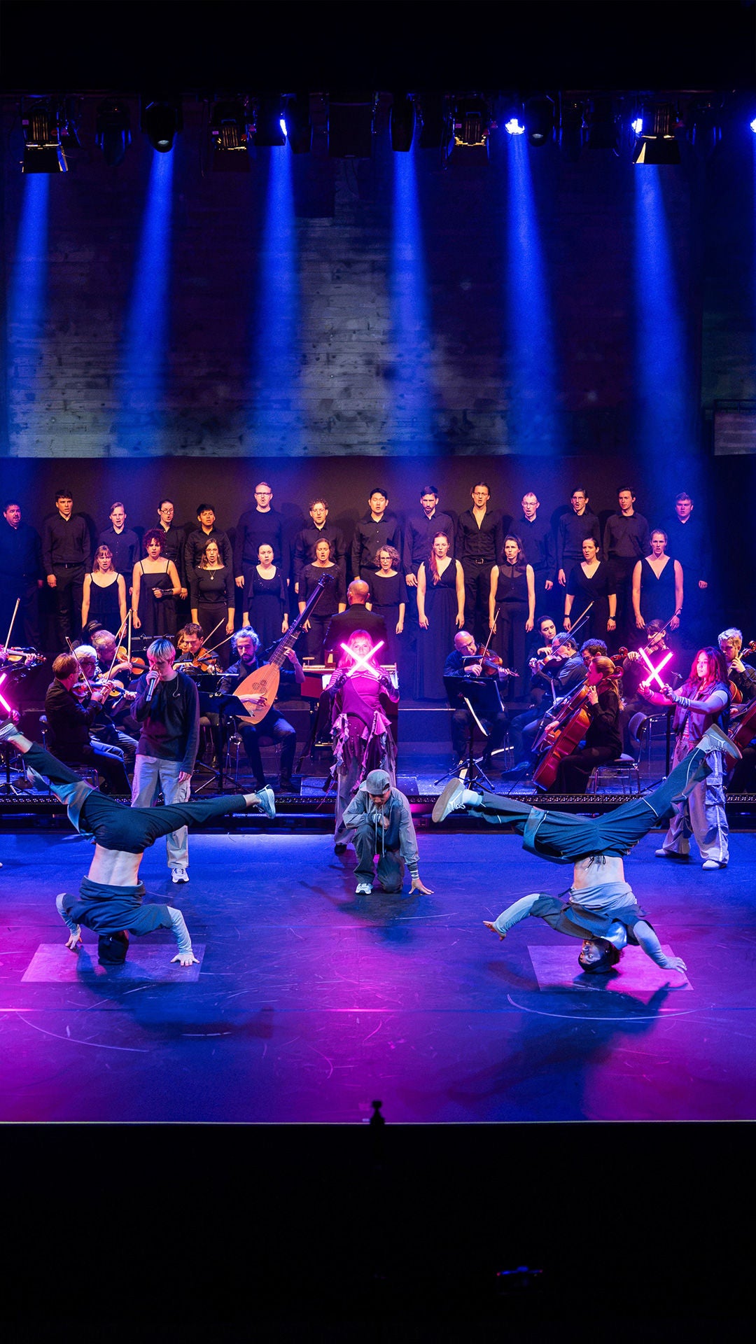Orchester mit Dirigent spielt in einer Konzerthalle. Die Bühne wird mit blauvioletten Lich bestrahlt. Vor dem Orchester bewegt sich eine Gruppe von drei break Dancern.