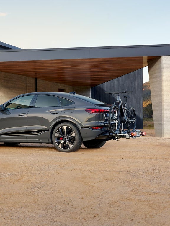 Grauer Audi Q6 Sportback e-tron mit Fahrrad am Heckträger vor moderner Garage auf sandigem Untergrund.