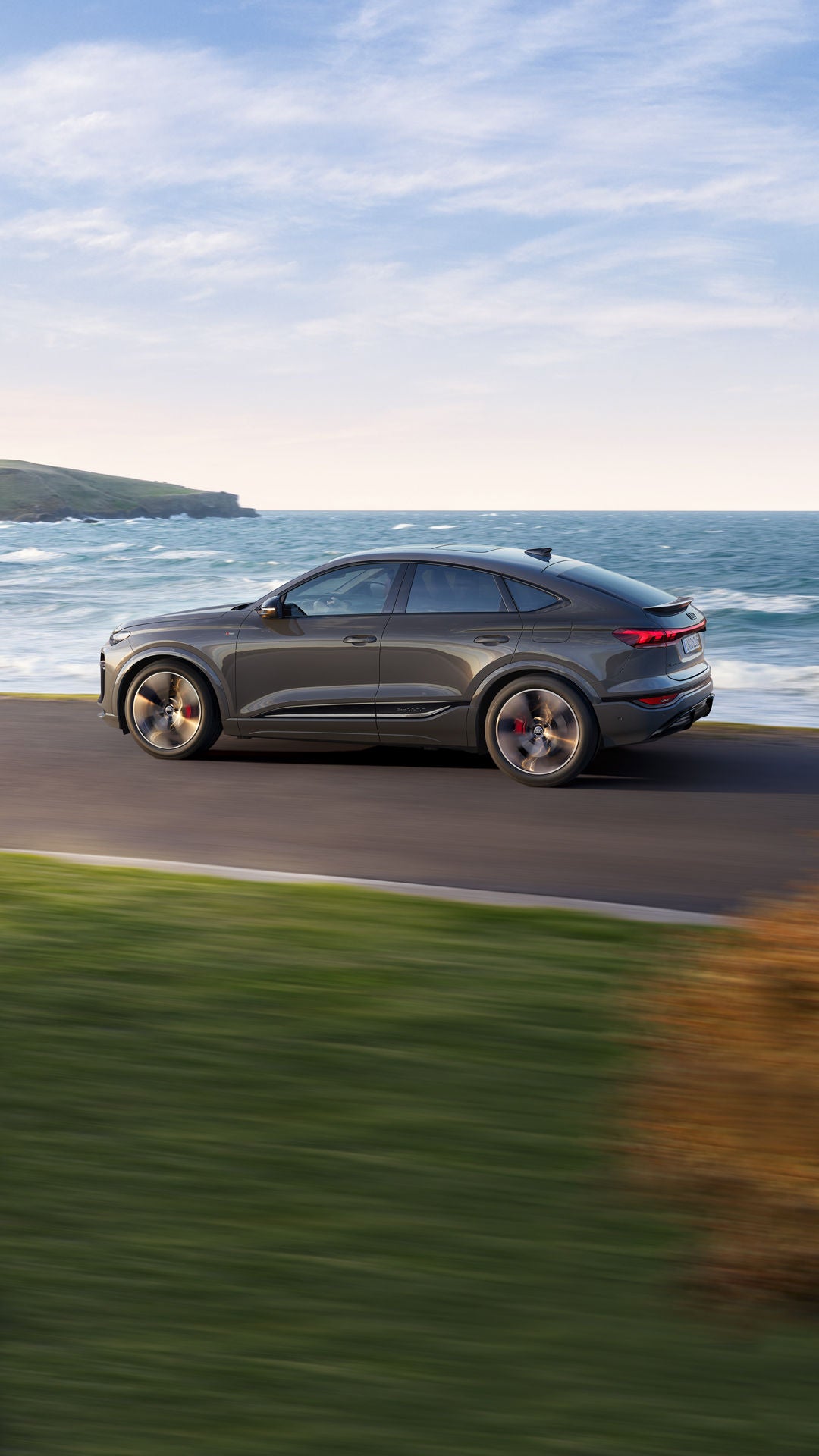 Seitliche Aufnahme des Q6 Sportback e-tron, welcher auf einer Straße entlang des Meeres fährt.