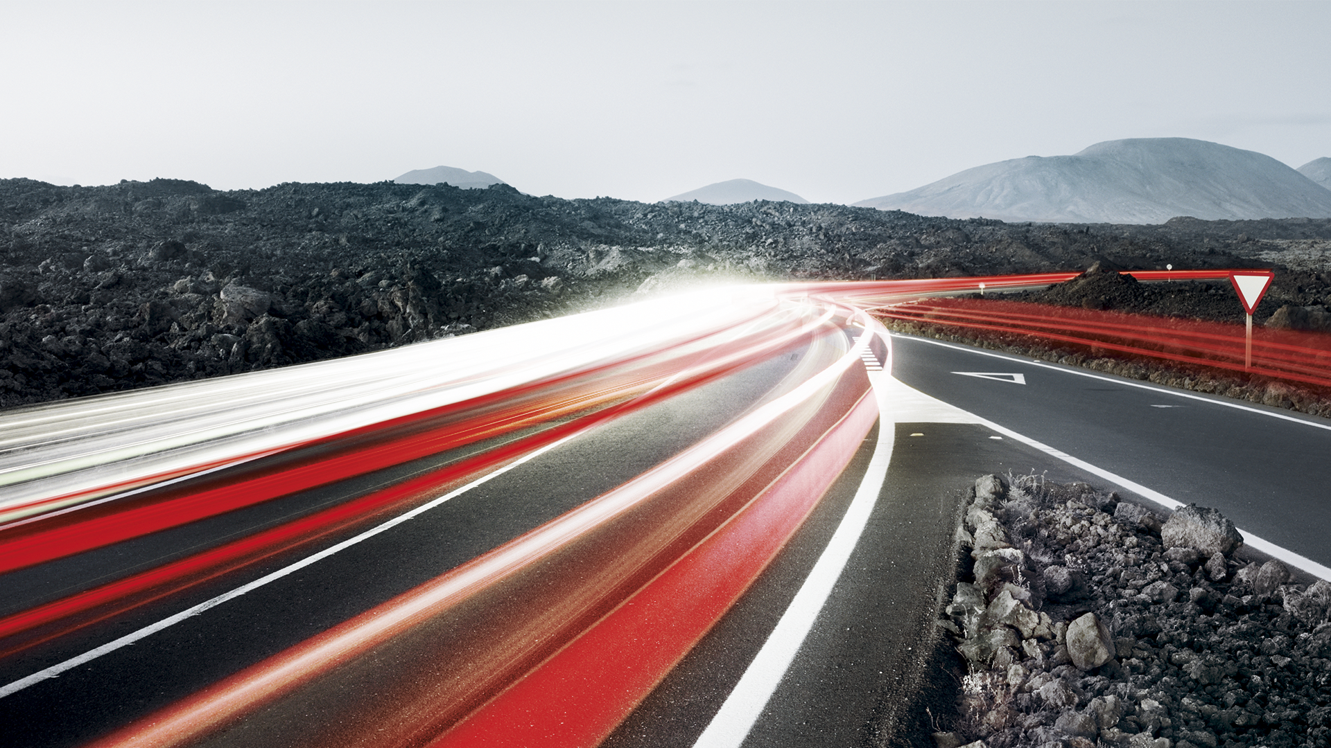 Lichtspuren von Autos auf kurviger Straße durch karge Vulkanlandschaft bei Dämmerung.