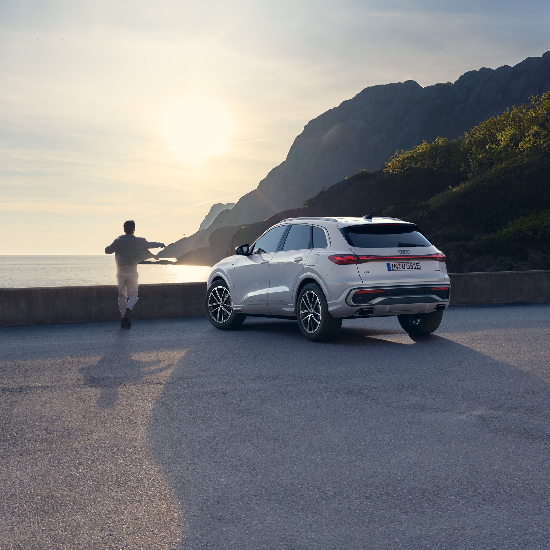 Eine Person steht an einer Küstenstraße mit Blick aufs Meer, daneben ist ein weißes Auto geparkt. Im Hintergrund ragen steile Felsen auf; die tief stehende Sonne wirft lange Schatten auf die Straße.