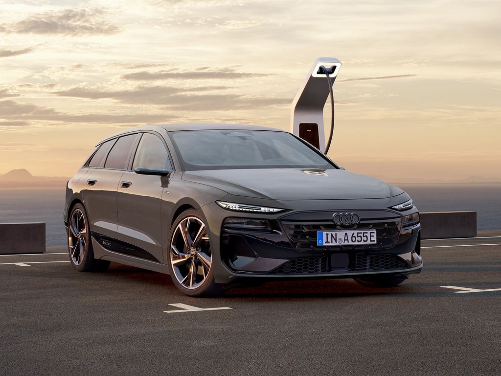 De Audi A6 Avant e-tron staat achterstevoren geparkeerd op een parkeerplaats en is aangesloten op een laadstation. Op de achtergrond zijn de zee en de bergen te zien.