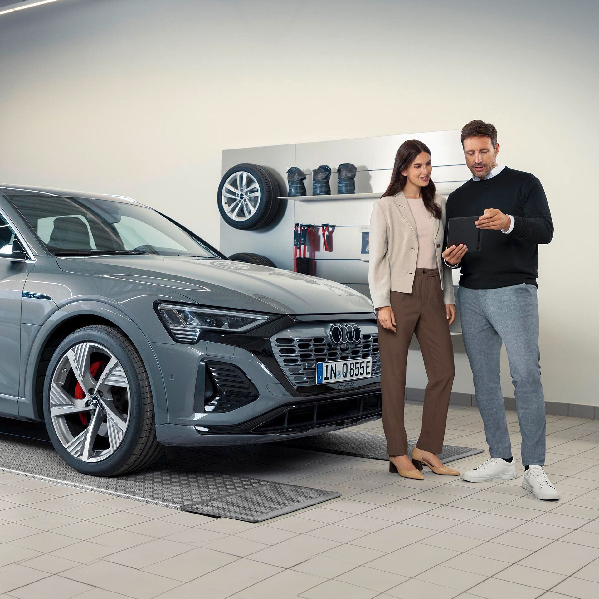 Mann zeigt Frau etwas auf einem Tablet neben einem grauen Audi in einer Werkstatt mit Felgen und Zubehör an der Wand.