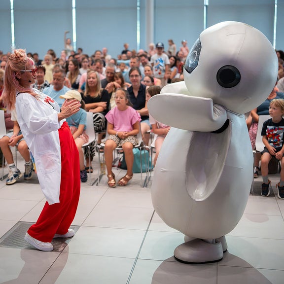Frau im weißen Kittel steht neben einem weißen humanoiden Roboter vor Publikum.