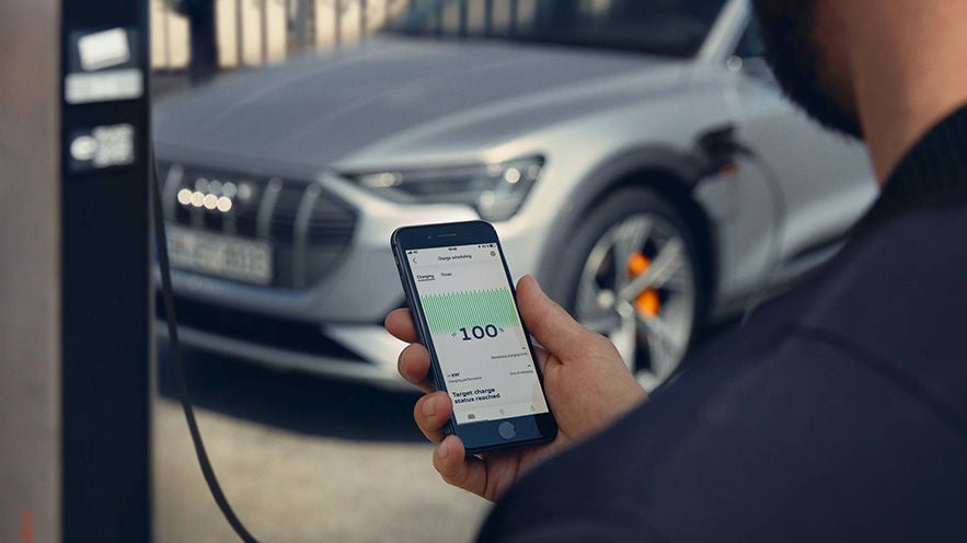 Person hält Smartphone mit Ladeanzeige bei 100 %, im Hintergrund steht ein silberner Audi e-tron an der Ladesäule.