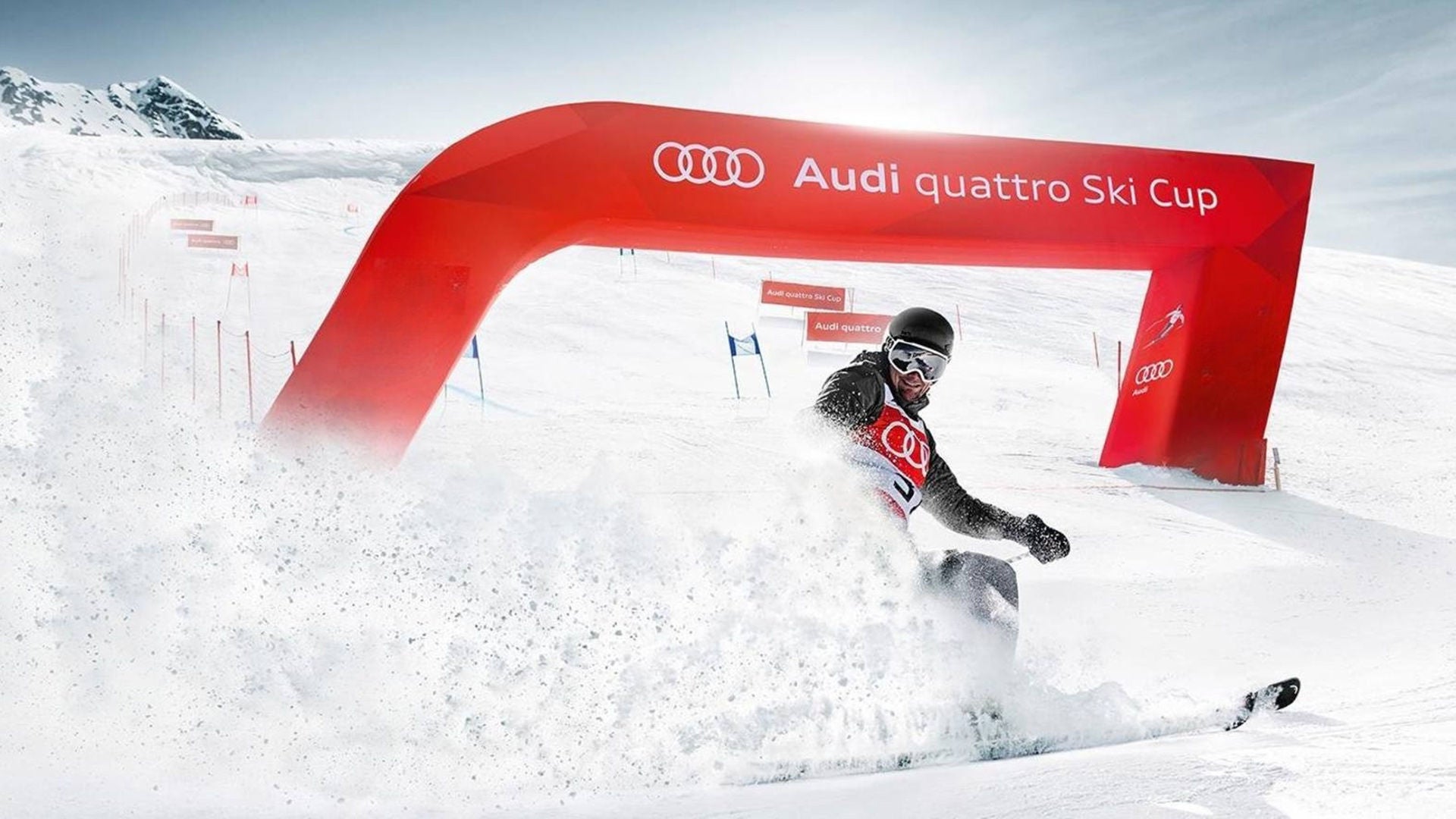 Skiers kick up snow in front of a red arch bearing the words “Audi quattro Ski Cup” on a snow-covered slope.
