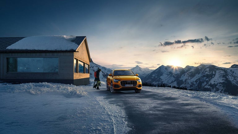 An orange Audi is parked in a mountainous landscape next to a hut and someone is on their way to the car