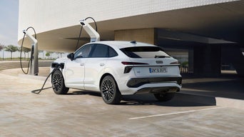 A white Q3 Sportback e-hybrid charges at a modern station, under a curved building with a sunny, open setting. The scene conveys innovation and sustainability.