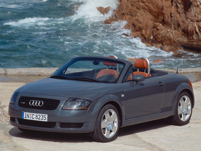 Front side view of a grey Audi TT Roadster quattro in front of rocks in the sea