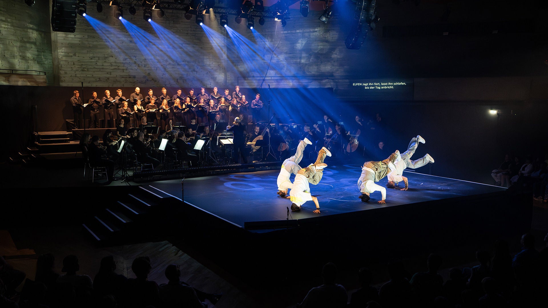 Breakdancer in weißen Outfits auf beleuchteter Bühne, im Hintergrund Chor und Orchester in Schwarz.