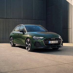 Dark green Audi parked in front of sleek, dark building. The car's modern design and angular headlights convey a sense of luxury and sophistication.