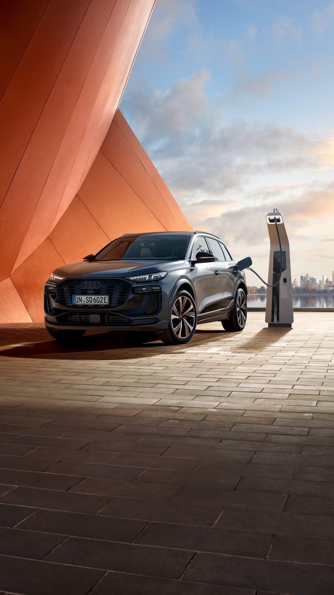 Seitliche Front des SQ6 SUV e-tron, welcher neben einer orangenen Gebäudefassade an eine Ladestation angeschlossen ist. Im Hintergrund ist das Meer, sowie die Skyline einer Stadt zu erkennen.