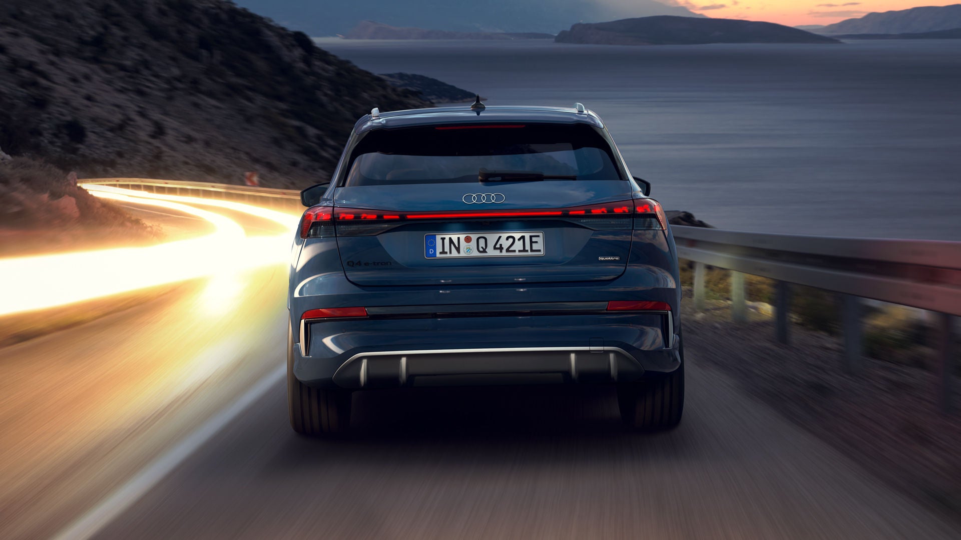 De Q4 SUV e-tron, ’s nachts van achteren gezien op een bochtige bergweg, met rechts de zee. Op de tegenliggende rijbaan zijn de koplampen van naderende voertuigen te zien. 