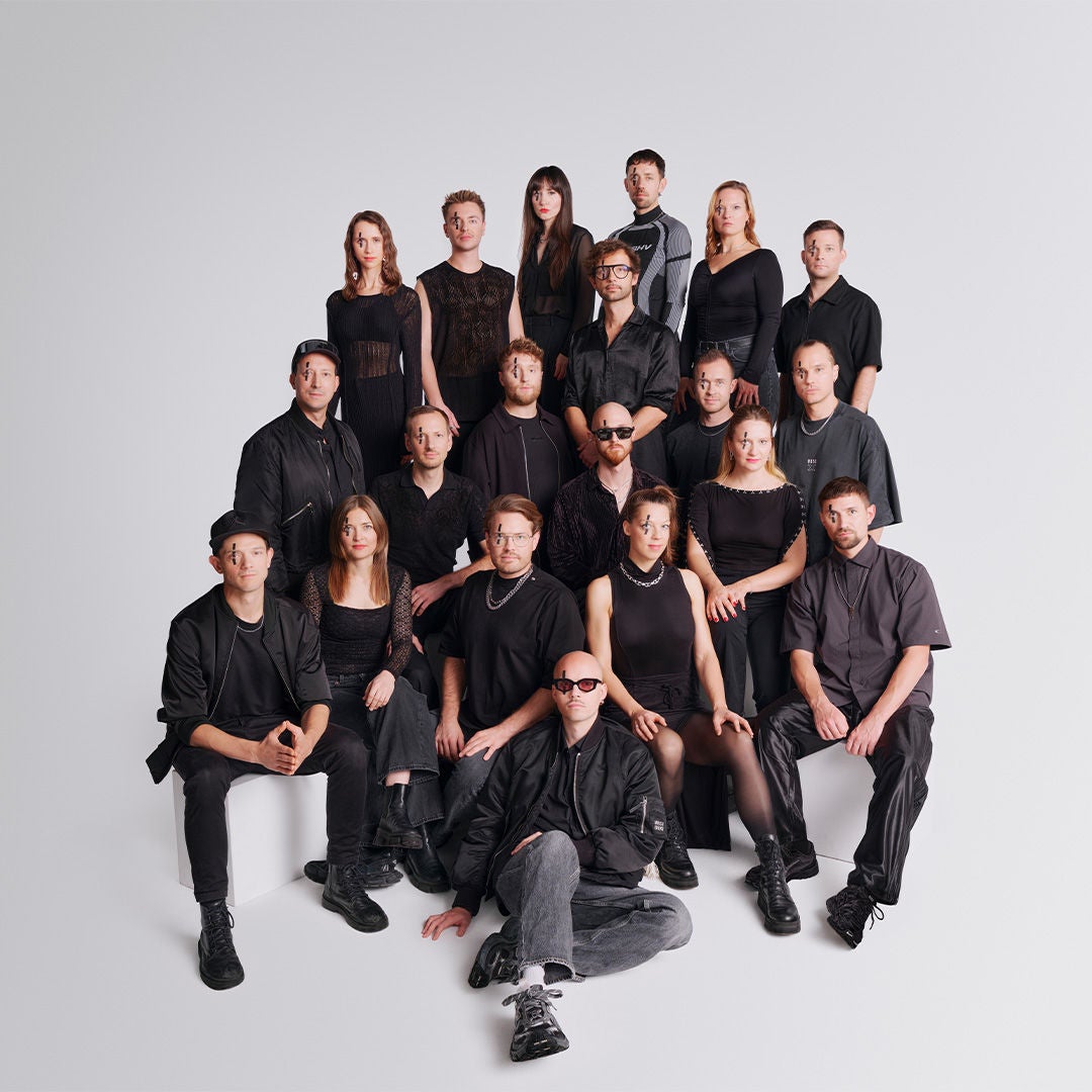 Gruppe von etwa 18 Personen in schwarzen Outfits mit auffälligem Gesichts-Make-up vor weißem Hintergrund.