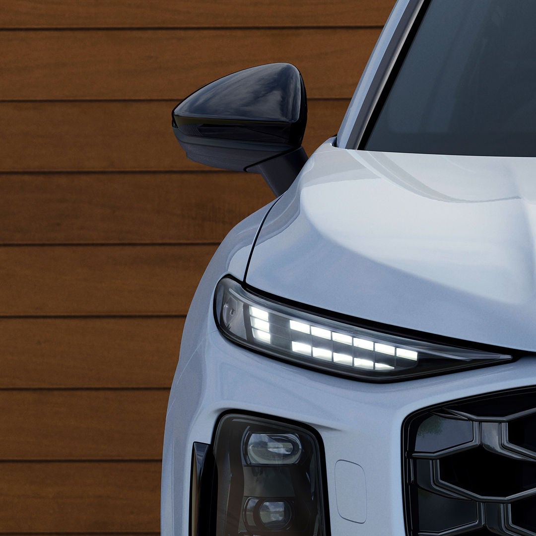 Close-up of the front left side of a white car against a wooden background. The sleek design features LED headlights and a glossy side mirror.