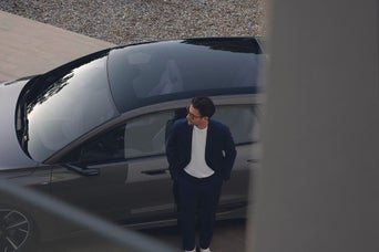Person im dunklen Anzug steht neben grauem Audi mit Glasdach, aufgenommen aus erhöhter Perspektive.