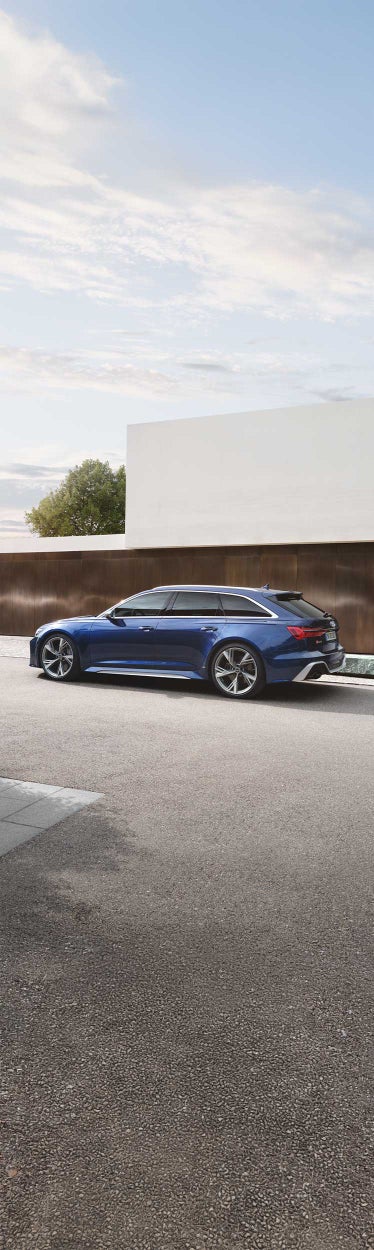 Seitliche Ansicht eines blauen Audi RS 6 Avant vor moderner Architektur, dunkelhaarige Frau im Businesslook in beigen Anzug steht daneben.