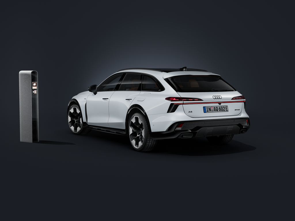 The Audi A6 allroad e-hybrid, viewed from an angle from behind against a dark background. Next to it is a modern charging point.