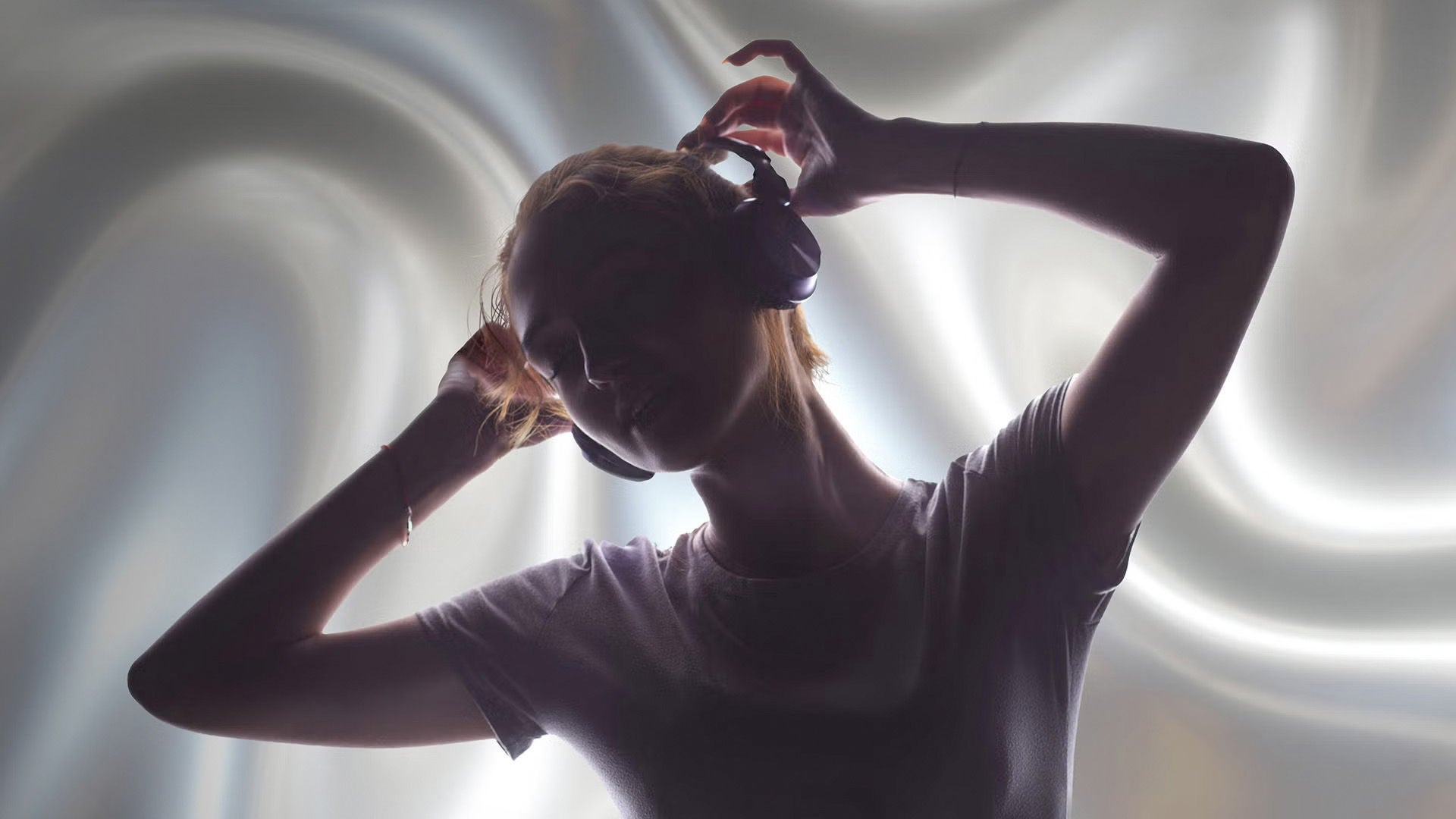 Person mit Kopfhörern im Profil hebt die Hände an den Kopfhörer, vor hellem, weich verschwommenem Hintergrund.