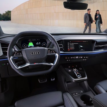 Cockpit des Audi Q4 e-tron mit digitalen Anzeigen, Touchscreen und Blick auf ein Paar draußen vor einer hellen Wand.