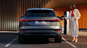 An Audi is parked and charging at a modern charging station, with a stylish individual handling the plug.