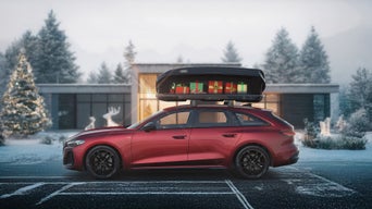 Ein roter Audi A6 Avant mit einer geöffneten Dachbox voller Weihnachtsgeschenke steht vor einem modernen Bungalow in verschneiter Winterlandschaft.