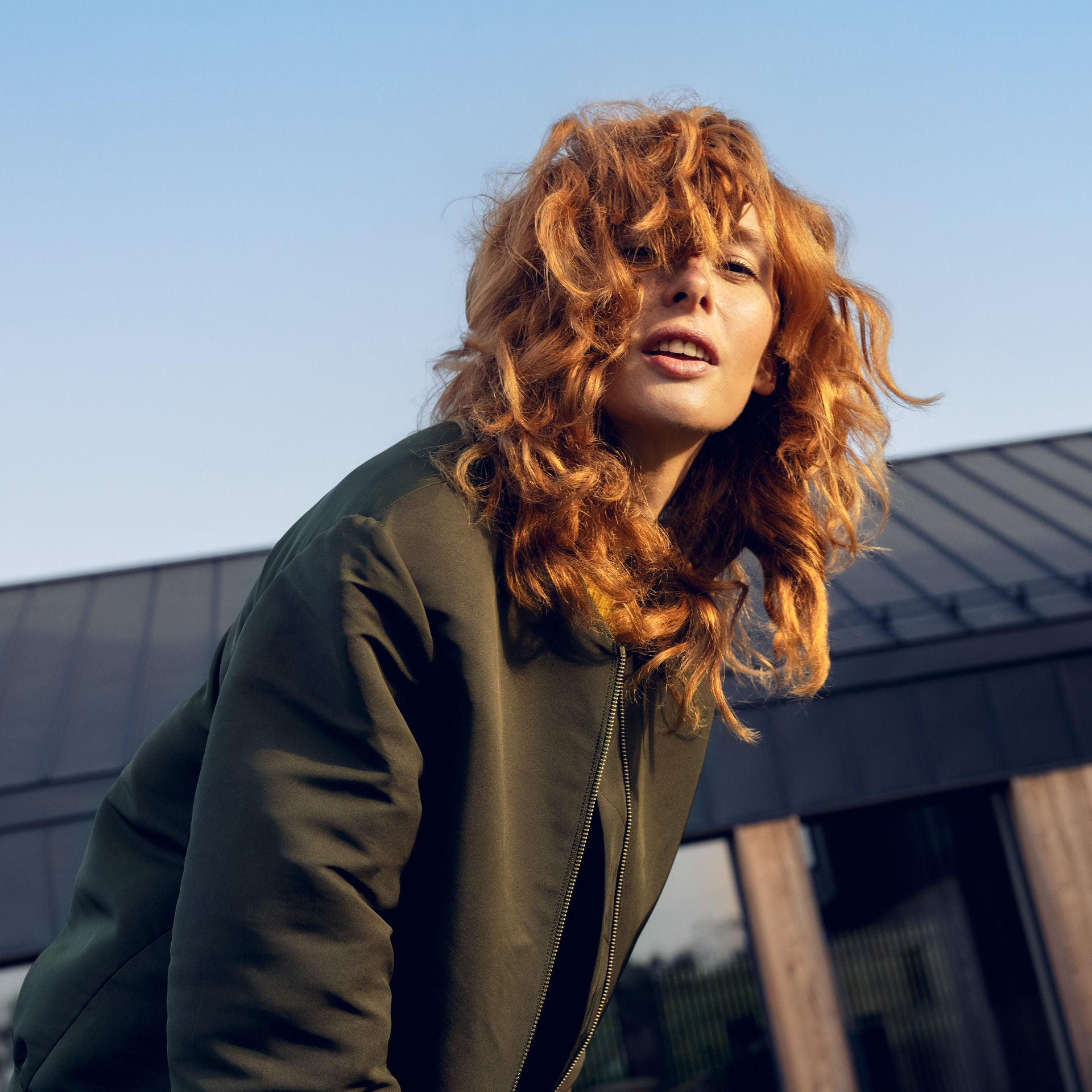 Frau mit lockigem, rötlichem Haar und grüner Jacke im Freien vor moderner Architektur bei Tageslicht.