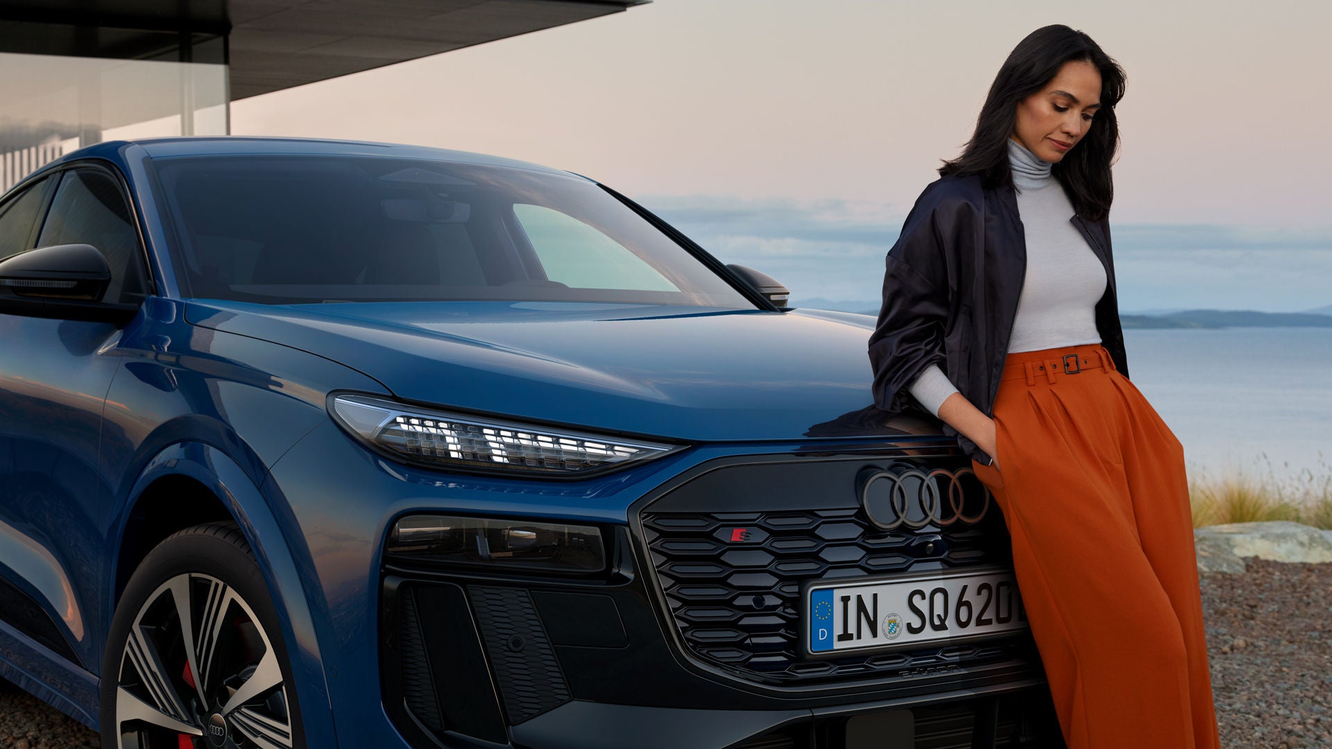 Schräges Frontprofil des SQ6 Sportback e-tron, welcher vor einem modernen Haus an der Küste steht. Eine junge Frau ist an die Motorhaube gelehnt.