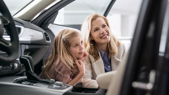 Lächelnde Frau mit kleinem Mädchen, beide sitzen nebeneinander im geöffneten Auto und schauen gemeinsam nach ins Auto.