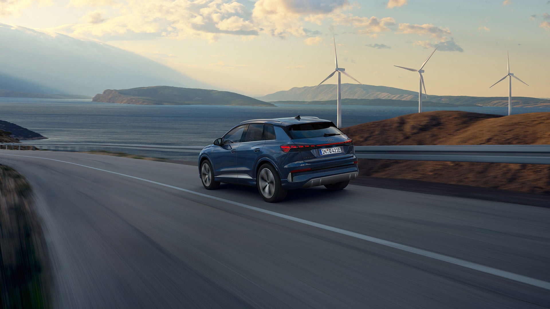Une Q4 e-tron bleue roule sur une route à grande circulation qui longe la mer. On aperçoit des éoliennes en arrière-plan.