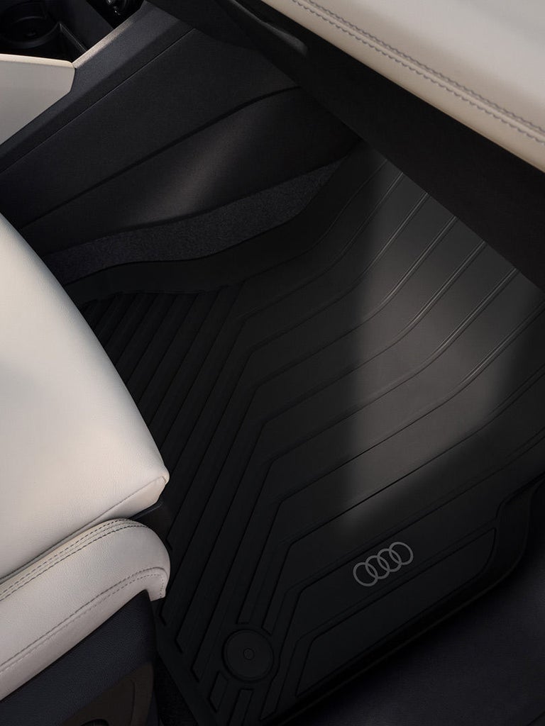 Detailansicht eines hellen Ledersitzes im Audi Q6 SUV e-tron mit schwarzer Gummifußmatte und Audi-Ringen im Fondbereich.
