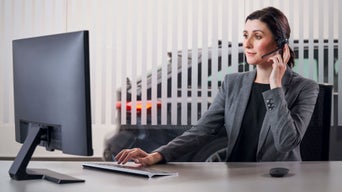 Eine Frau im grauen Blazer telefoniert und schaut auf einen Computerbildschirm