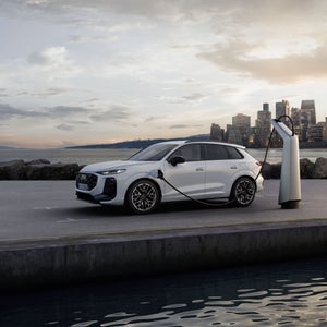 De Q3 e-hybrid staat gekoppeld aan een laadpaal met op de achtergrond de skyline van een stad.